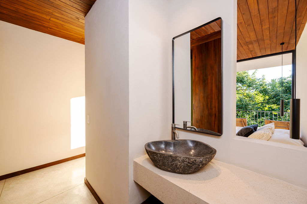 Stone basin on a cast concrete shelf beneath a black-framed mirror, open to the bedroom beyond